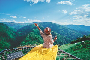 young free carefree girl traveler wearing hat relaxing with glass wine sitting hammock during enjoying happy traveling moment life against background green big mountains